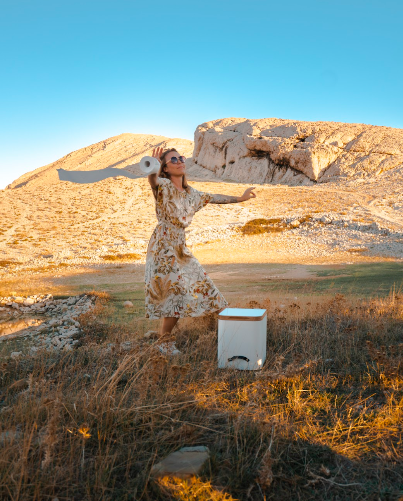 Frau tanzt um die Trenntoilette Timber herum auf der Wiese, Berge im Hintergrund, Frau hält Toilettenpapier in der Hand.