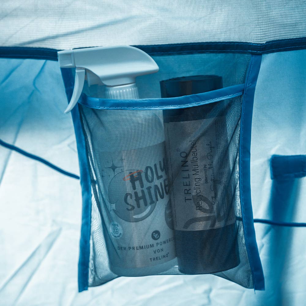 Blaues Netzfach mit zwei Holy Shine Sprayflasche und Müllbeutel im Inneren eines Toilettenzelts.