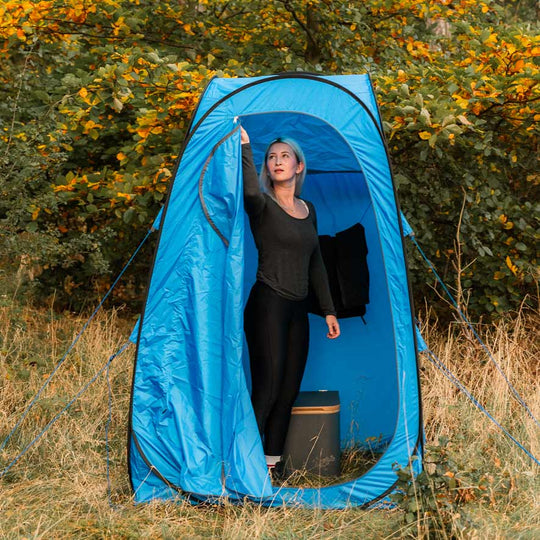 Blaues Popup-Toilettezelt mit Reißverschluss, Frau öffnet Vorhang, Waldhintergrund.